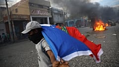 Ein vermummter Mann trägt eine Haiti-Flagge, Im Hintergrund brennen Reifen