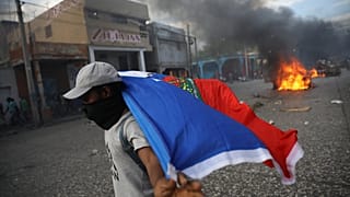Ein vermummter Mann trägt eine Haiti-Flagge, Im Hintergrund brennen Reifen