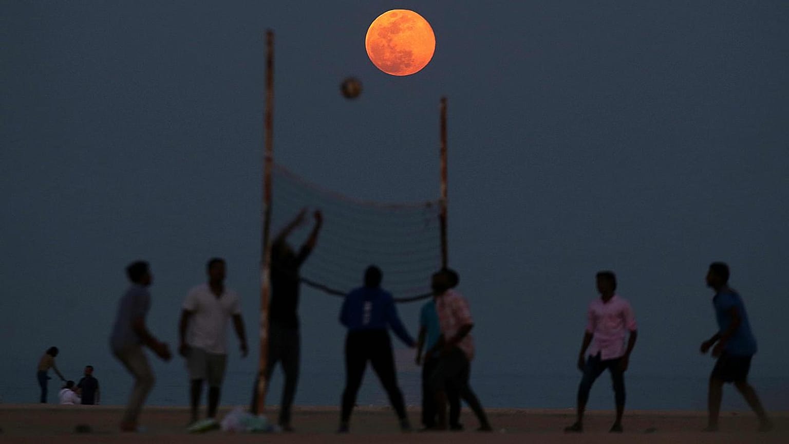 People play volleyball on the beach as the Pink Moon rises in the sky over 