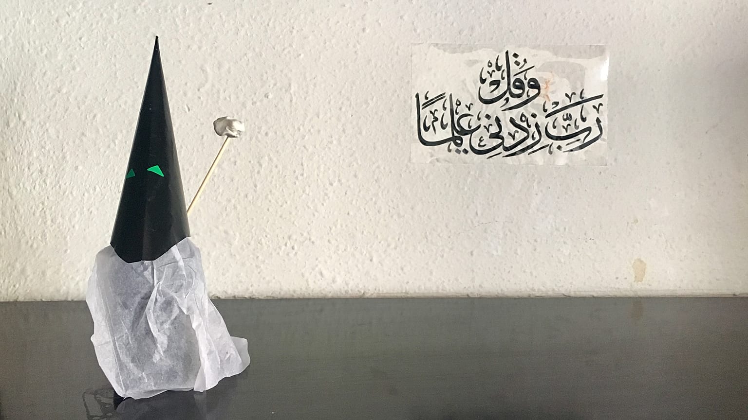 Capirote de Semana Santa hecho con papel.Al fondo, una inscripción coránica