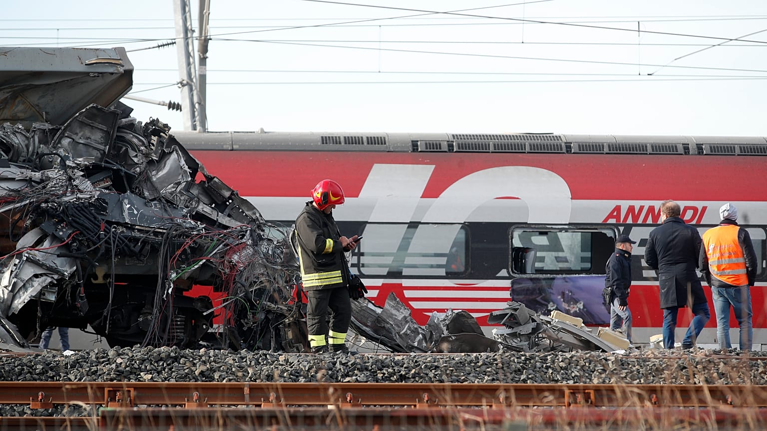 The wreckage of the locomotive of a high-speed train crammed onto another train on adjacent tracks, as the rest of train is seen in the background, after it derailed. Italy.