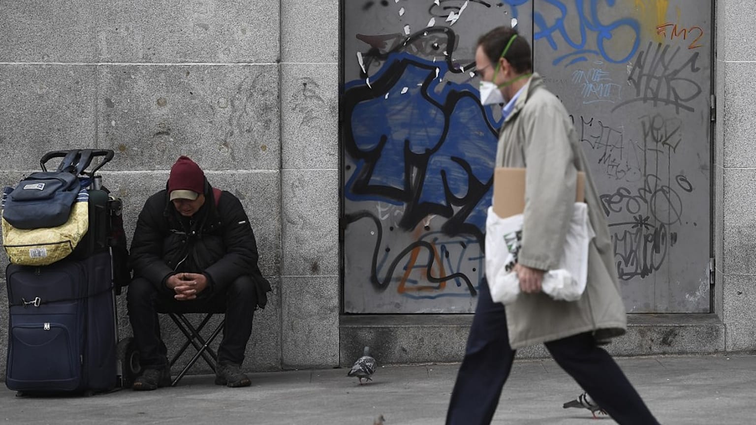 Un hombre protegido con una máscara pasea cerca de un 'sin techo' en Puerta del Sol, Madrid