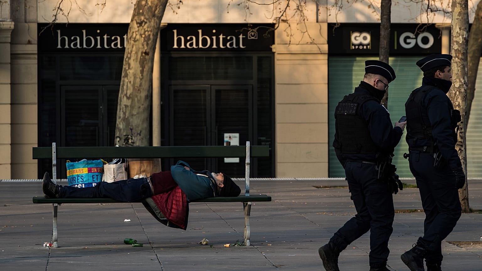 Policemen walks by a homeless man sleeping on a bench on March 17, 2020 in Paris