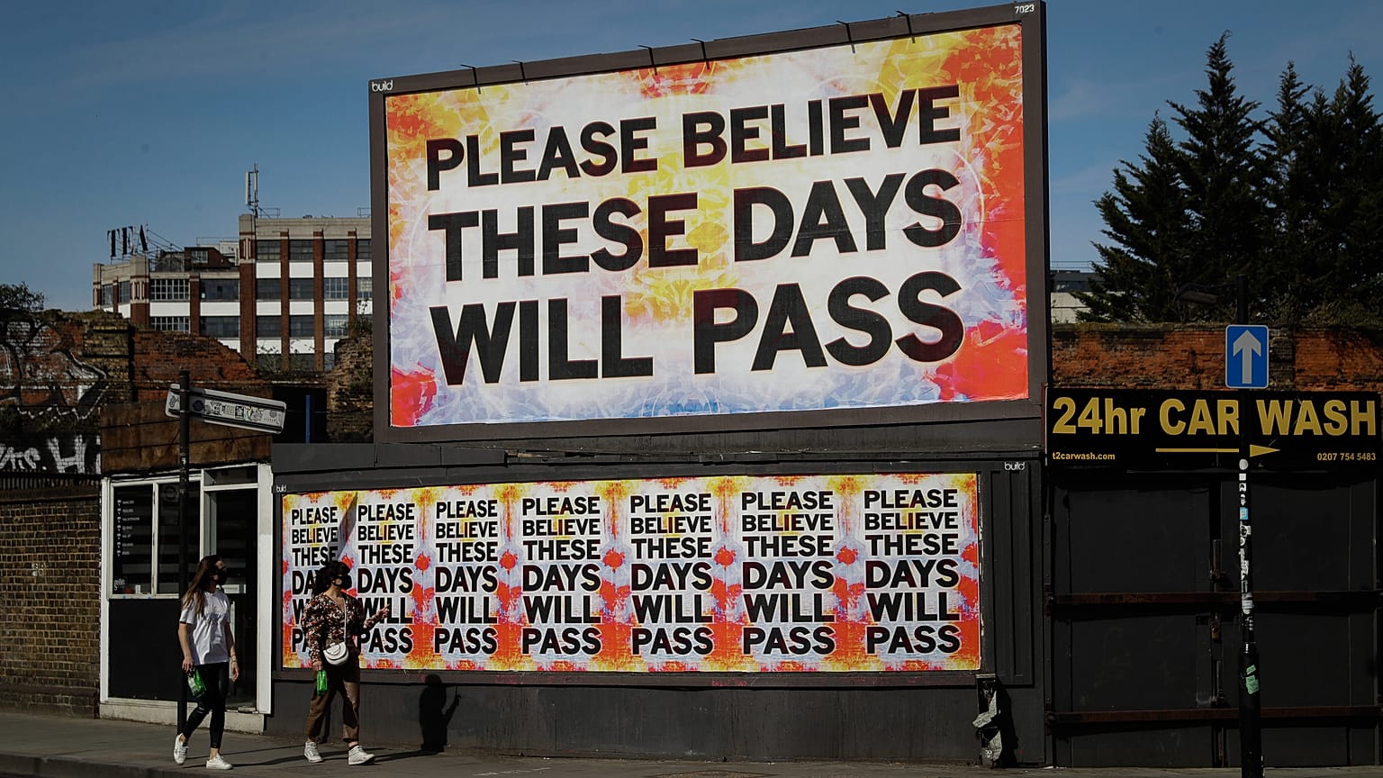Women wearing face masks to protect from coronavirus walk past a billboard with a work by artist Mark Titchner in east London