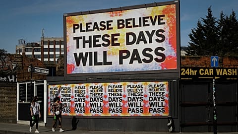 Women wearing face masks to protect from coronavirus walk past a billboard with a work by artist Mark Titchner in east London