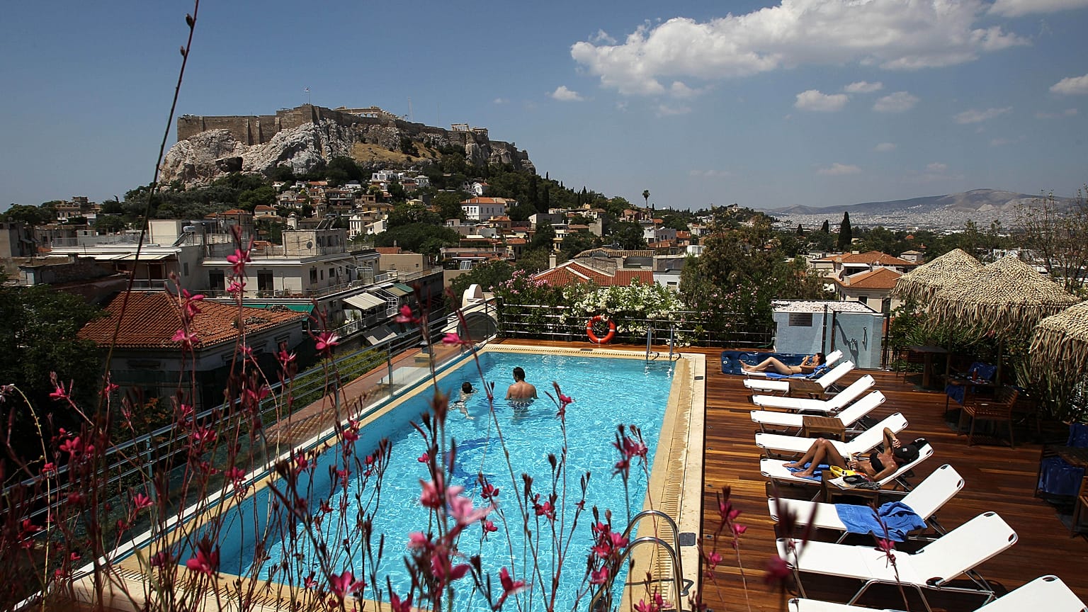 Tourists swim in a swimming pool on the roof of a hotel as the ancient Acropolis hill 