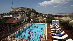 Tourists swim in a swimming pool on the roof of a hotel as the ancient Acropolis hill 