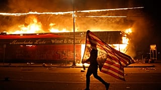 Un hombre con una bandera estadounidense frente a un comercio en llamas en Mineápolis