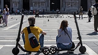 A woman and a man relax sitting on their scooters at the Duomo square, in Milan, Italy, Monday, May 25, 2020.
