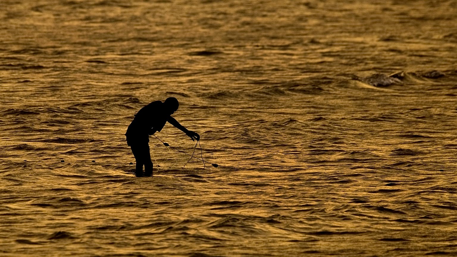A fisherman standing in the ocean adjusts his nets at sunset on the shoreline of Djibouti city, Djibouti