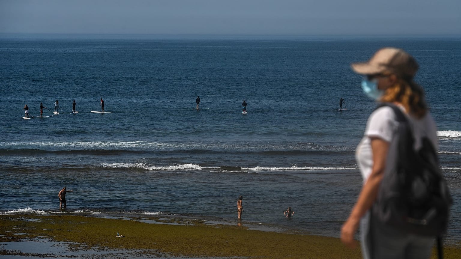 A woman wearing a face mask walks past paddle surfers riding waves at Parede beach in Cascais in the outskirts of Lisbon