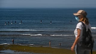 A woman wearing a face mask walks past paddle surfers riding waves at Parede beach in Cascais in the outskirts of Lisbon
