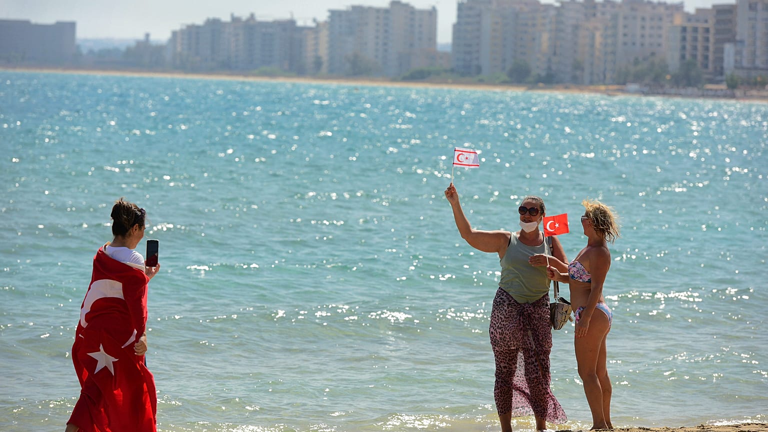 زوار يحملون الأعلام التركية المنفصلة يلتقطون صورًا لبعضهم البعض على الشاطئ مع المباني المهجورة بعد أن فتحت الشرطة شاطئ فاروشا.