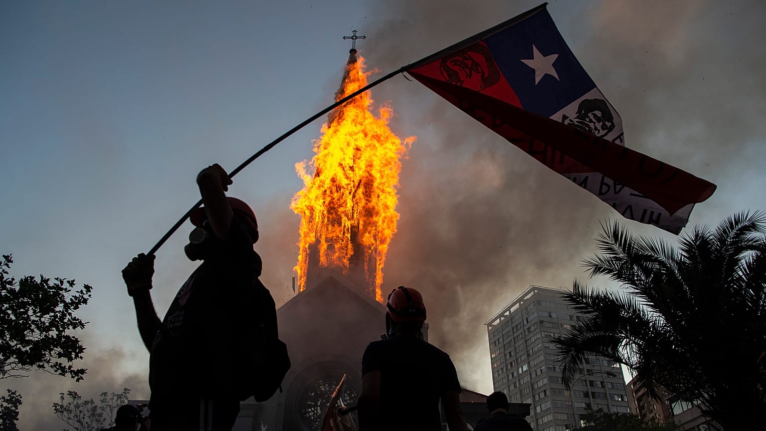 Un manifestante enarbola una bandera de Chile frente a la cúpula de la Asunción en llamas