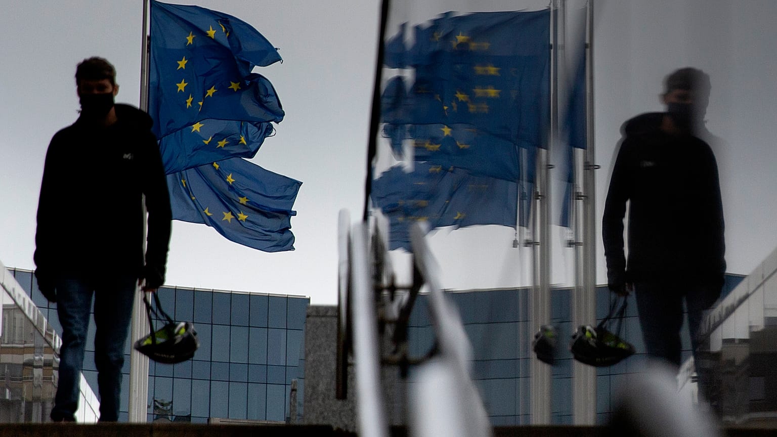 A man carries a cycling helmet as he walks by EU flags outside EU headquarters in Brussels, Wednesday, Oct. 28, 2020.