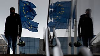 A man carries a cycling helmet as he walks by EU flags outside EU headquarters in Brussels, Wednesday, Oct. 28, 2020.