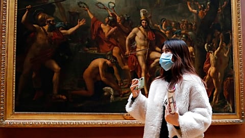 Masked tourist walks by an oil on canvas of 1814 entitled Leonidas at Thermopylae, by Jacques Louis David, at the Louvre Museum in Paris