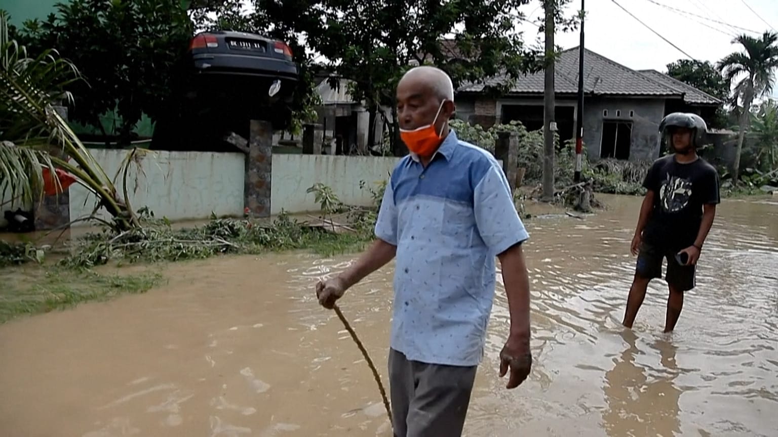 Men walking on muddy road past the car swept away by the flood sitting above a fence.