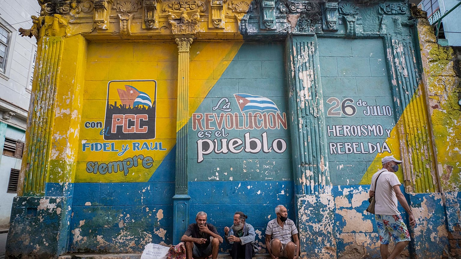 Tres hombres con las mascarillas mal colocadas charlan sentandos delante de un muro con consignas políticas en La Habana