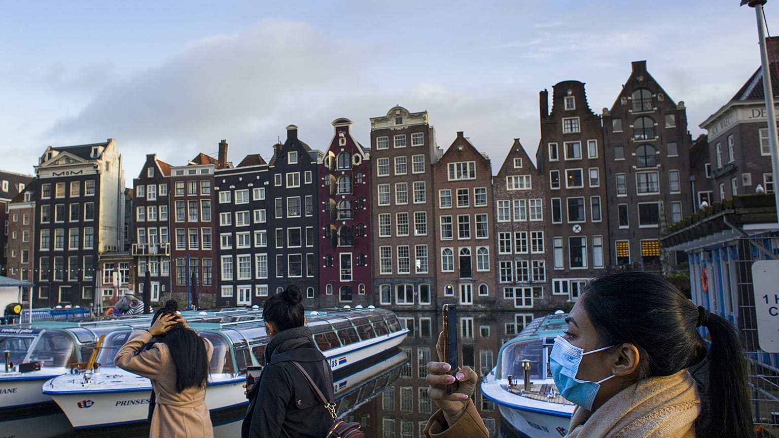 A woman, wearing a face mask to curb the spread of the COVID-19 virus, takes images of empty and moored canal cruise boats docked in Amsterdam, Netherlands, Thursday, Feb. 4, 