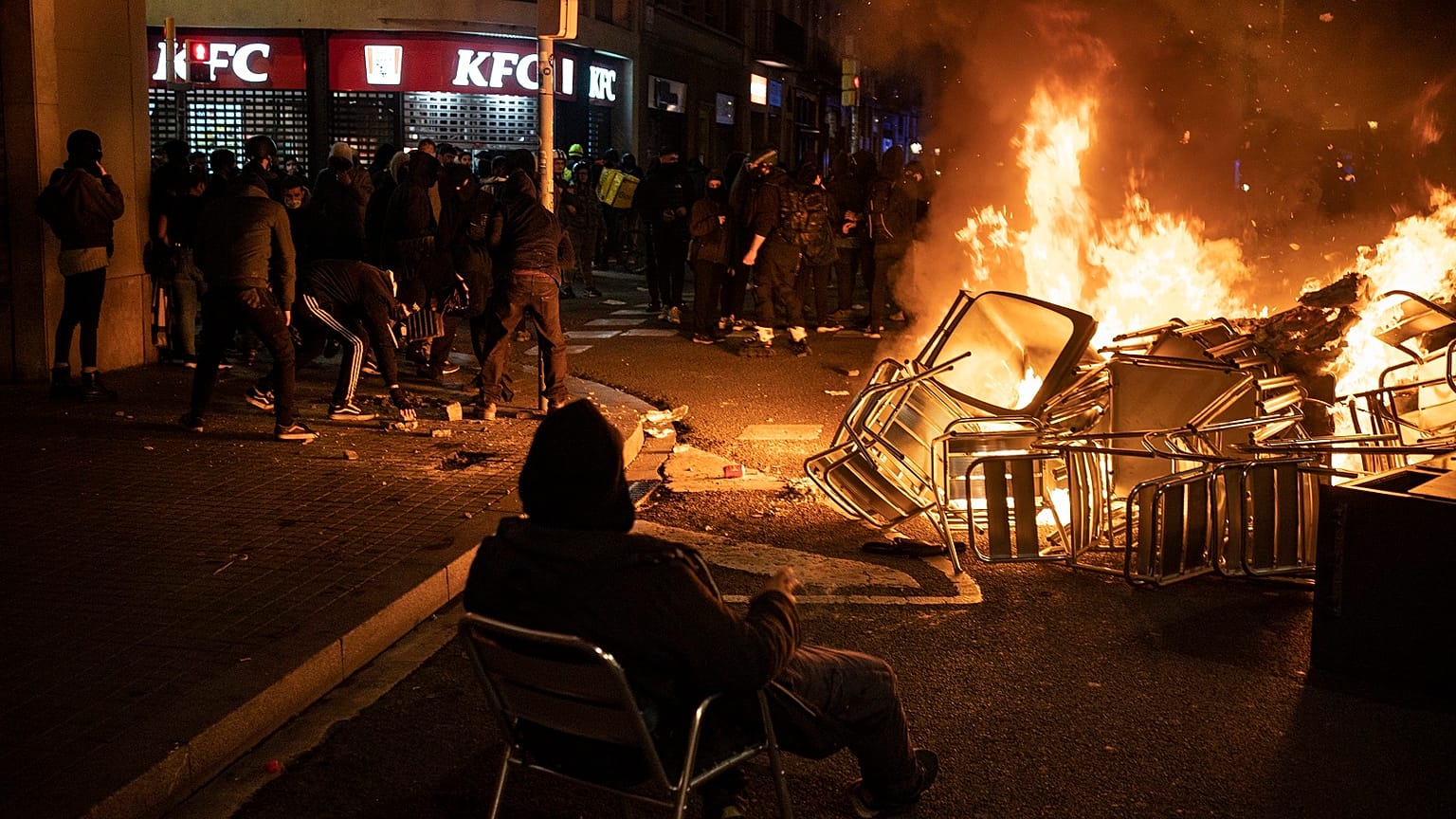 A man sits on a chair in front of a burning barricade during a protest condemning the arrest of rap singer Pablo Hasél in Barcelona, Spain, Friday, Feb. 19, 2021.