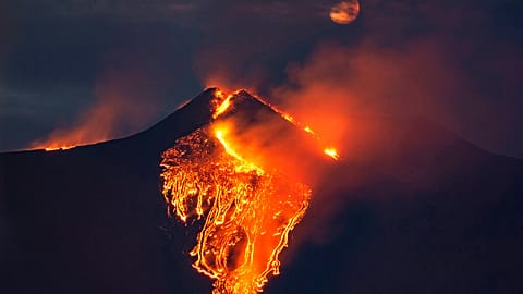 The moon is partially seen in the sky as lava flows from the Mt Etna volcano, near Catania in Sicily, southern Italy, early Tuesday, Feb. 23