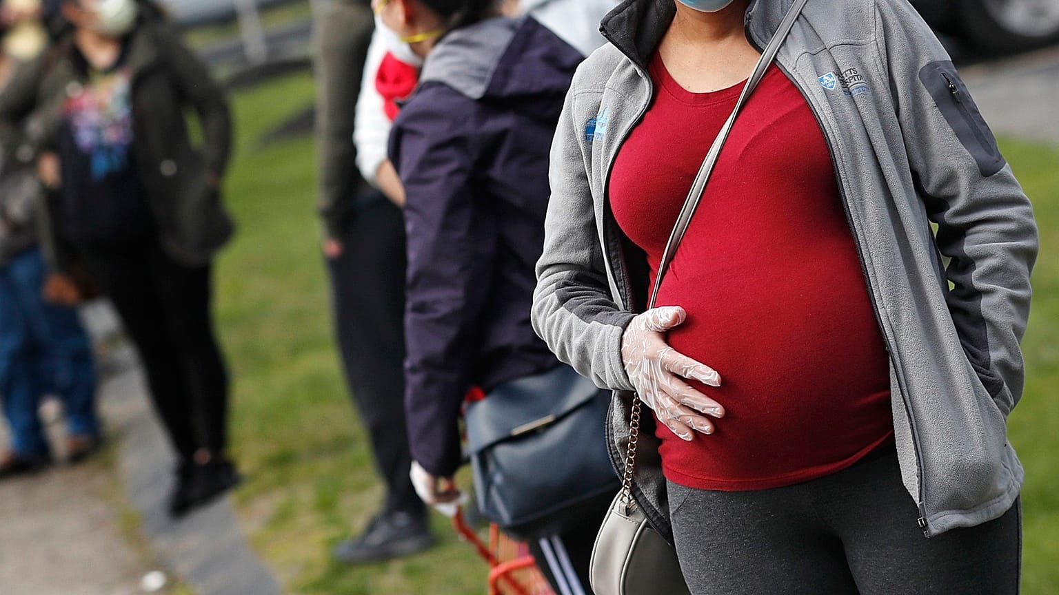 A pregnant woman wearing a face mask and gloves as precautions against COVID holds her belly.