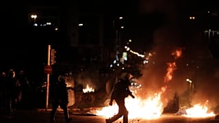 Αστυνομικός των ΜΑΤ περπατάει μπροστά από φωτιές 