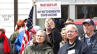 A sign denouncing the COVID-19 vaccine is held up in the crowd gathered at a protest