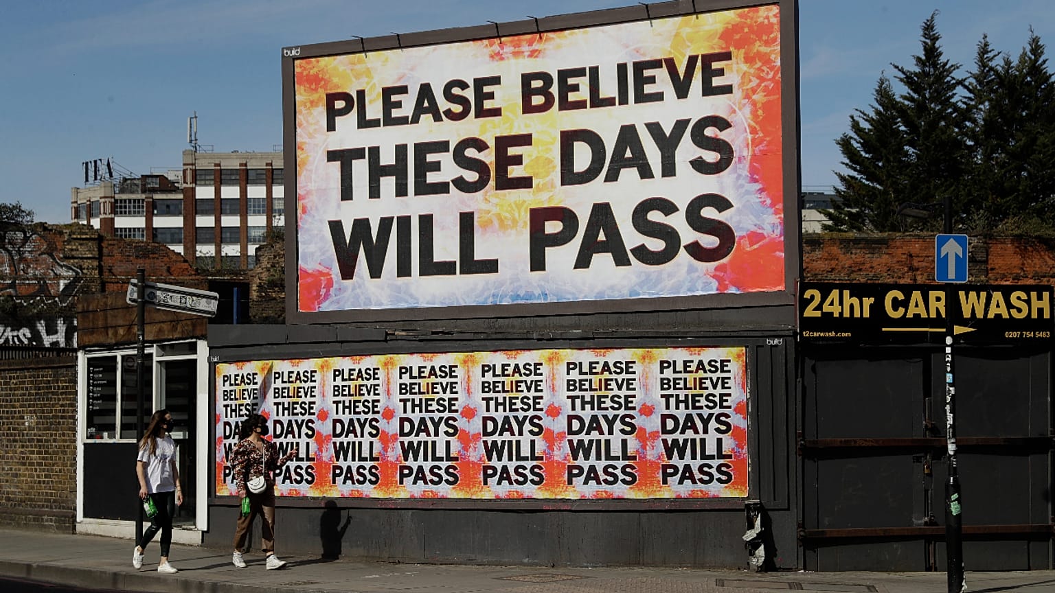 Women wearing face masks to protect from coronavirus walk past a billboard with a work by artist Mark Titchner in east London, Tuesday, April 7, 2020. 