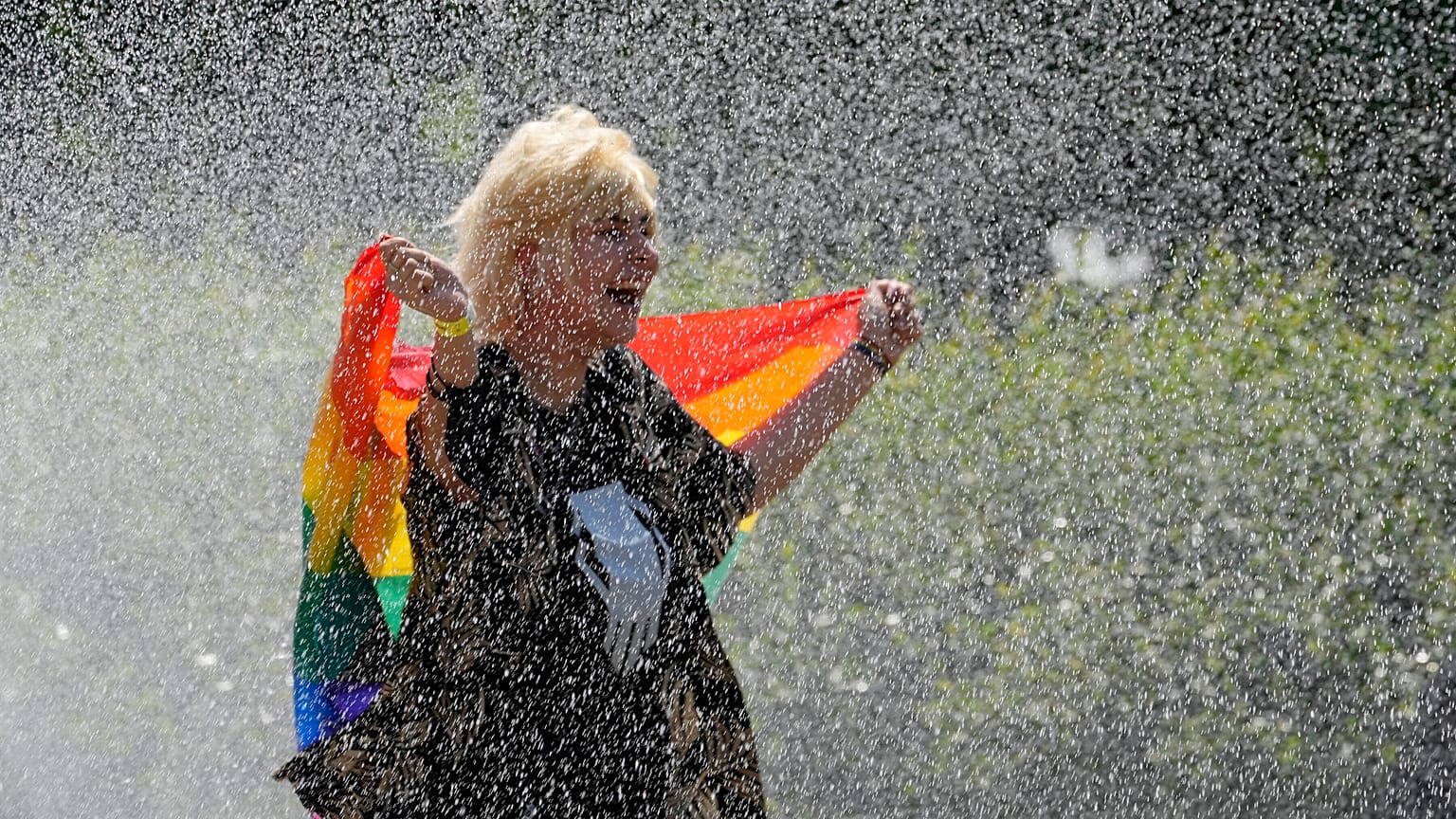A woman with a rainbow flag cools off in a sprinkler ahead of the Equality Parade, the largest LGBT pride parade. Warsaw, Poland. June 19, 2021