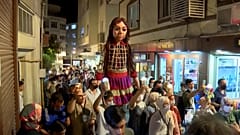 'Little Amal' walking down streets, greeting residents.