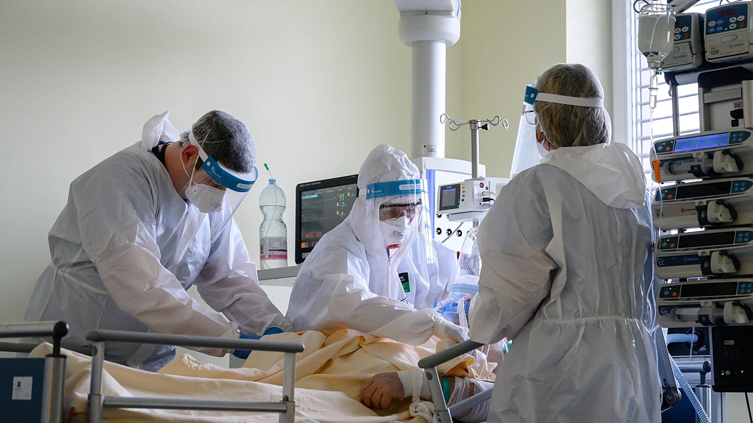 Védőfelszerelést viselő orvos és ápolók egy oxigénellátást segítő helmet sisakot helyeznek fel egy betegre 