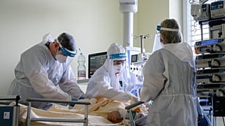Védőfelszerelést viselő orvos és ápolók egy oxigénellátást segítő helmet sisakot helyeznek fel egy betegre 