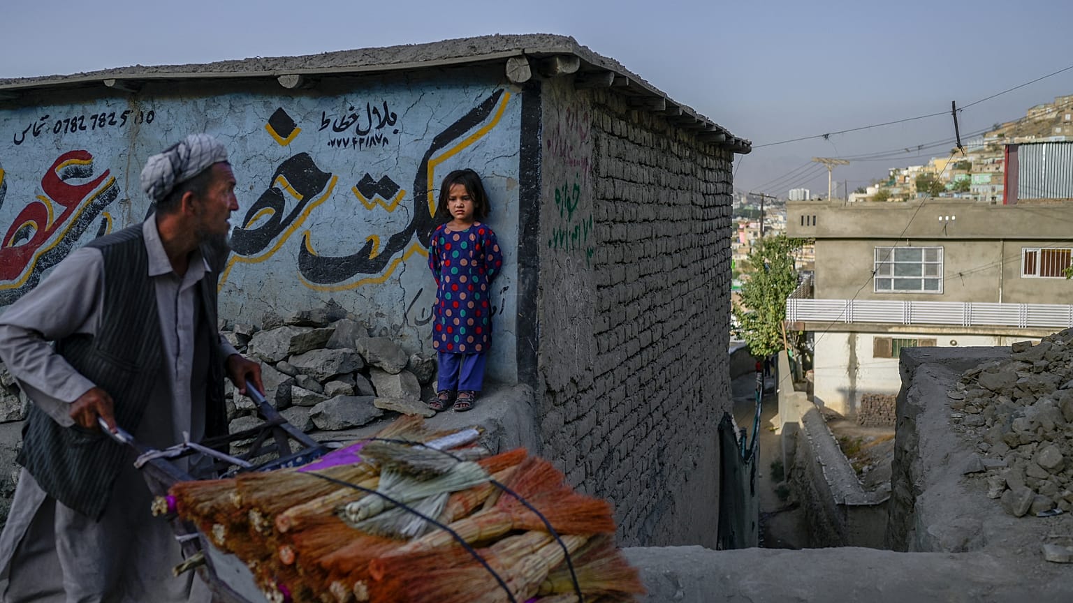 A vendor pushes his handcart loaded with brooms as a girl watches in Kabul.