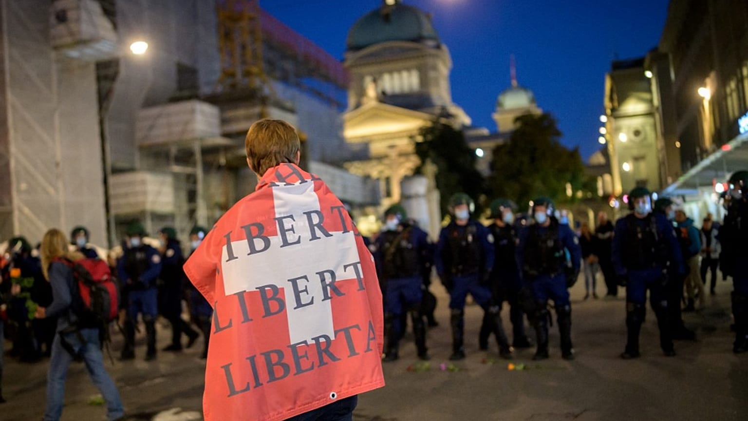 A man holding a Swiss flag on his shoulders reading "Freedom" walks past riot police.