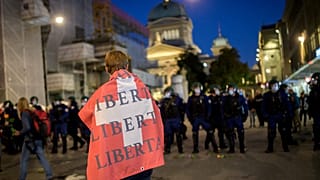 A man holding a Swiss flag on his shoulders reading "Freedom" walks past riot police.