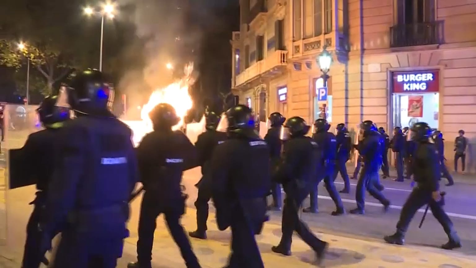 Un grupo de agentes antidisturbios avanza hacia una zona en la que varios individuos están quemando contenedores