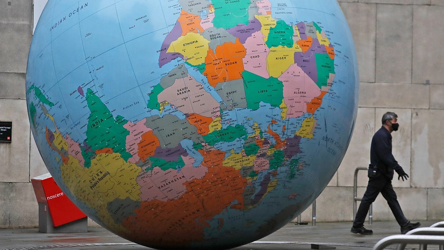 A man wearing a face mask walks past a globe in front of the London School of Economics in London, Tuesday, Jan. 19, 2021. An art piece called "The World Turned Upside Down".
