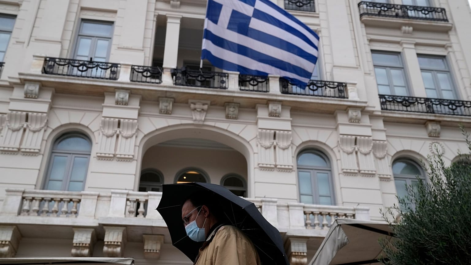 A man wearing a face mask to protect against coronavirus, walks outside a store as a Greek flag waves in Athens, Greece.