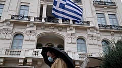 A man wearing a face mask to protect against coronavirus, walks outside a store as a Greek flag waves in Athens, Greece.