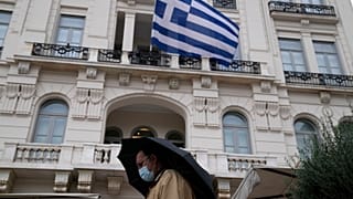 A man wearing a face mask to protect against coronavirus, walks outside a store as a Greek flag waves in Athens, Greece.