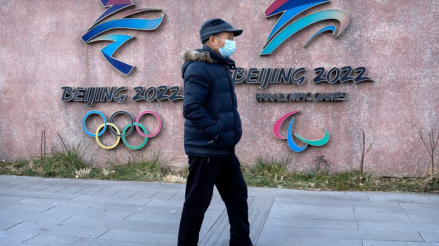 A man wearing a face mask walks past the logos for the Beijing 2022 Winter Olympics and Paralympics in Beijing, Thursday, Dec. 16, 2021.