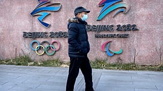 A man wearing a face mask walks past the logos for the Beijing 2022 Winter Olympics and Paralympics in Beijing, Thursday, Dec. 16, 2021.