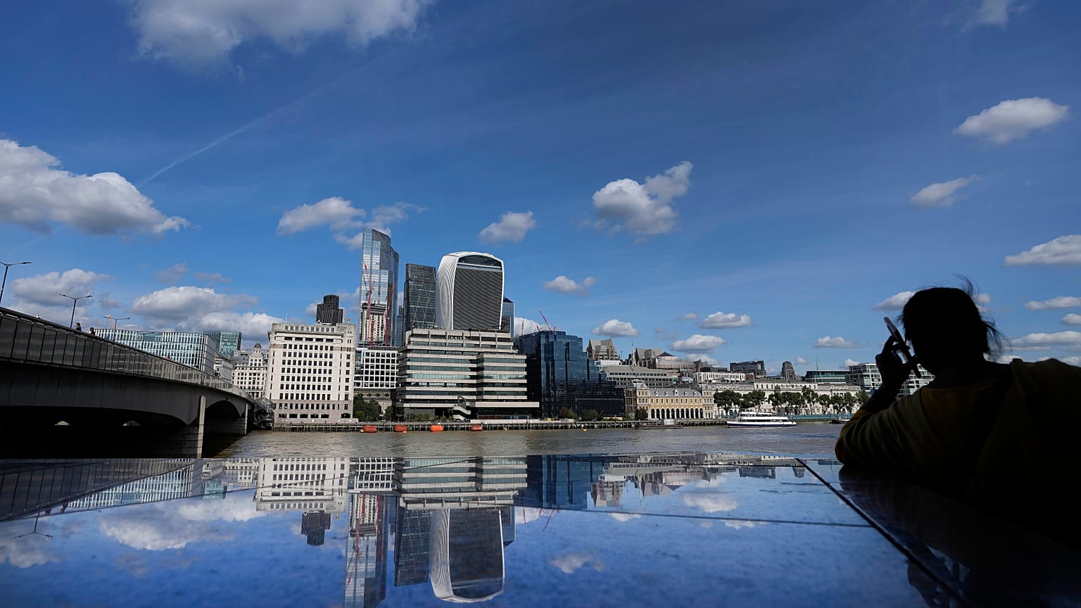 A woman is silhouetted as she takes a phone call, as the skyline of London is reflected in the polished stones of London Bridge during sunny weather in London, Tuesday, Sept. 