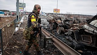 An armed man stands by the remains of a Russian military vehicle in Bucha, close to the capital Kyiv, Ukraine, Tuesday, March 1, 2022.