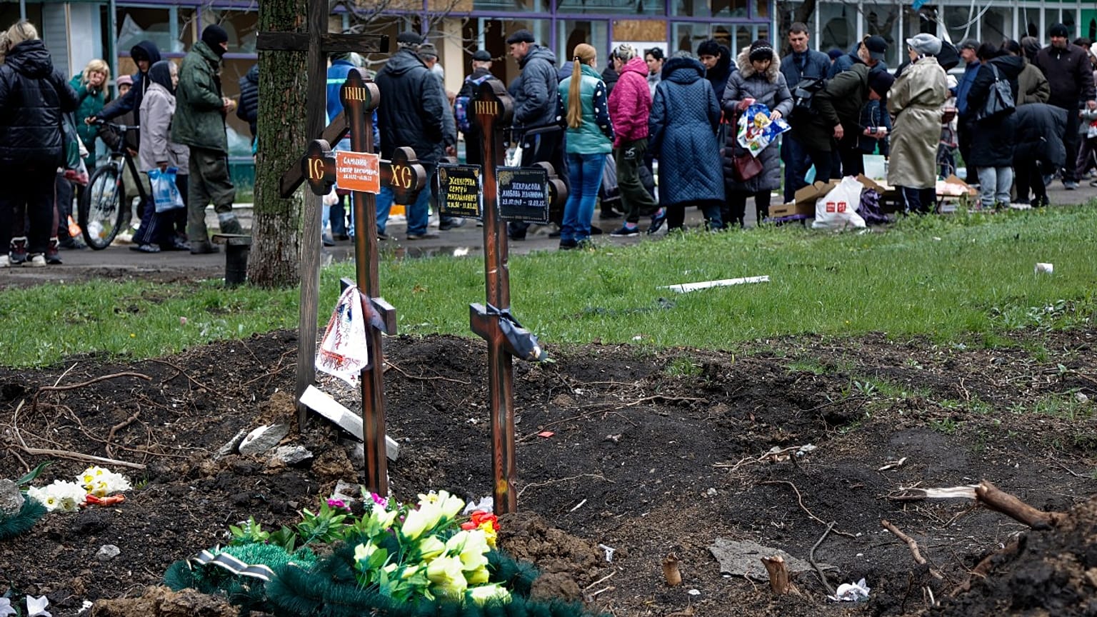 Menschen stehen Schlange auf einem behelfsmäßigen Straßenmarkt neben frischen Gräbern in Mariupol.