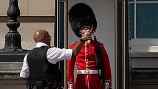 A police officer givers water to a British soldier wearing a traditional bearskin hat, on guard duty outside Buckingham Palace on 18 July 2022