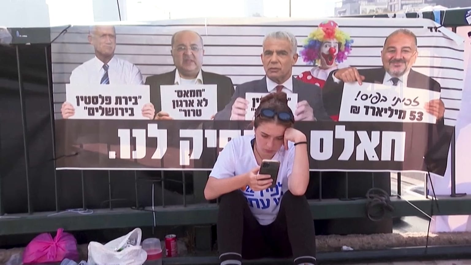 Una joven mira su teléfono m´óvil sentada frente a un cartel de la oposición a Netanyahu