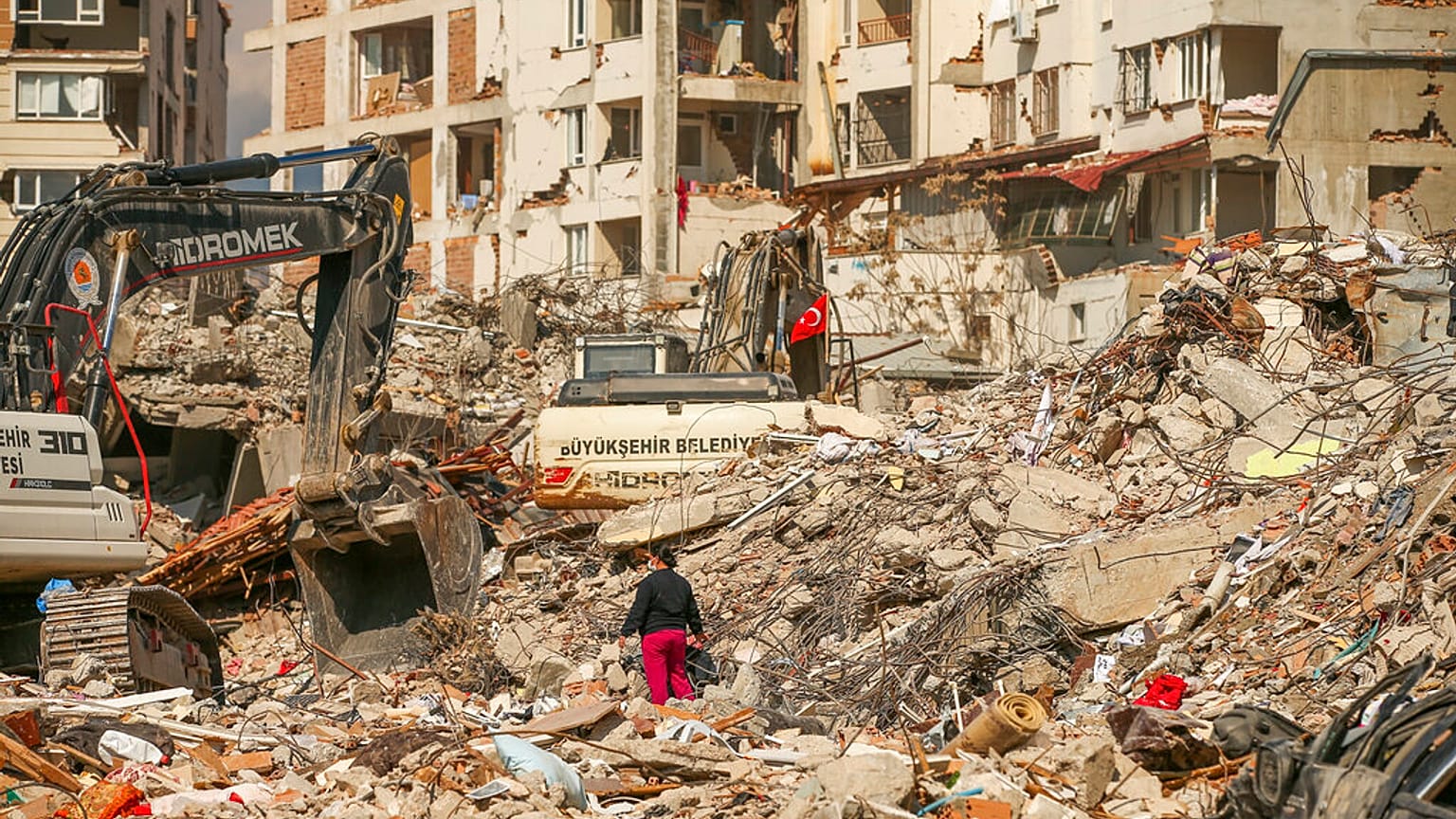 رجل يسير بين الأنقاض بينما تهدم الحفارات بقايا مبنى مدمر في أعقاب الزلزال الذي ضرب سمانداغ جنوب تركيا، 22 فبراير 2023.
